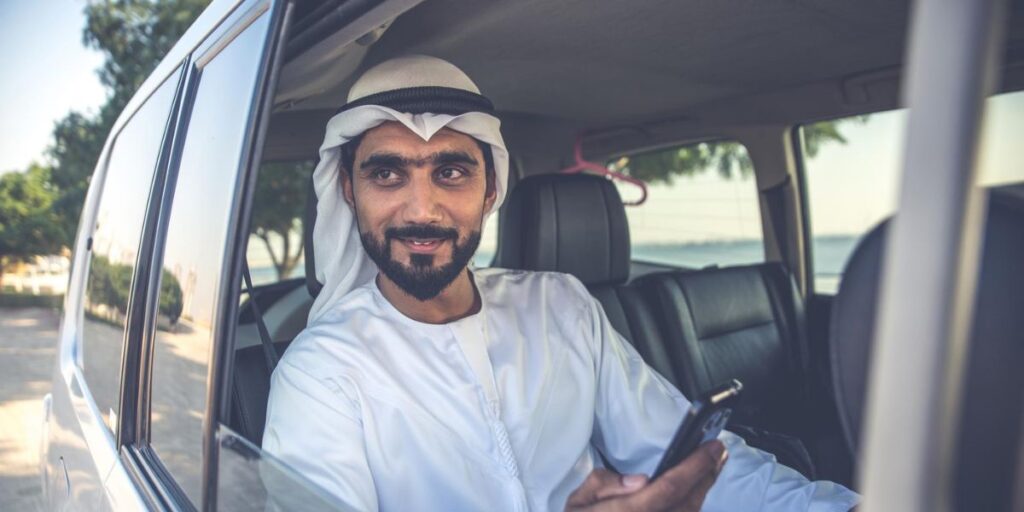A bearded man in a white thobe and ghutra sits in the back of a car while holding a smartphone and looking outside