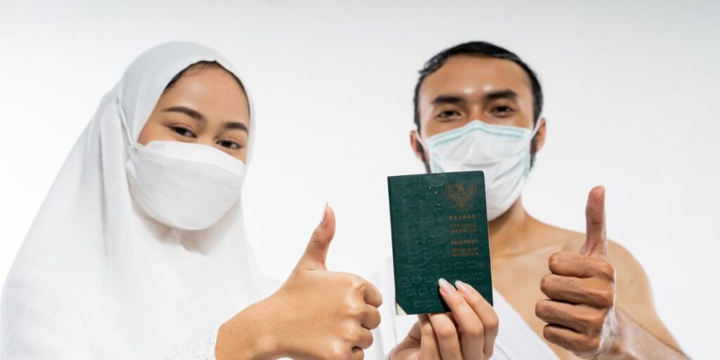 A man and woman in ihram wearing masks hold up an Indonesian passport and give a thumbs-up against a white background