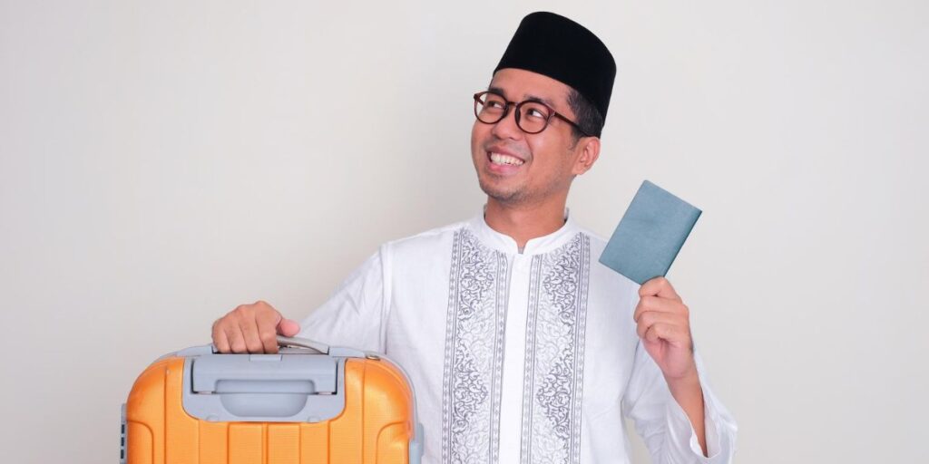 A smiling man in a songkok and white shirt holds an orange suitcase and his passport against a plain background
