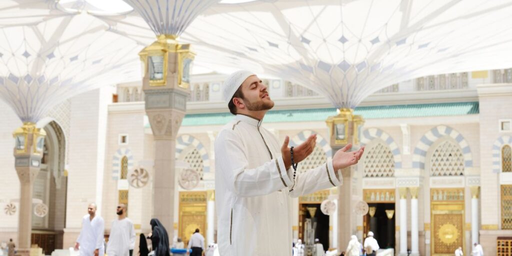 A man in white prayer clothes stands with open palms in the marble courtyard of the Prophet's Mosque in Medina
